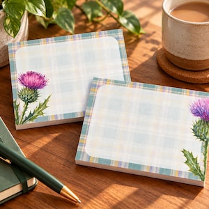 May include: Two rectangular sticky note pads featuring a light blue and white plaid border with a vibrant purple thistle flower illustration on the side. The paper has a smooth white centre area for writing notes, showcasing a botanical design.