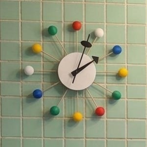 Peut inclure: Une horloge murale de style rétro avec un cadran blanc et des aiguilles noires. L'horloge comporte des marqueurs sphériques colorés en rouge, jaune, vert, bleu et blanc. L'horloge est fixée sur un mur carrelé vert clair.