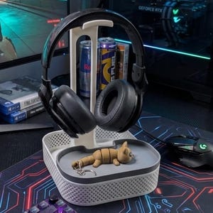 May include: A black gaming headset rests on a white and gray headphone stand, with a small wooden cat figurine in the tray below. Two cans of energy drink and a book are also in the stand. A black mouse and keyboard are visible.