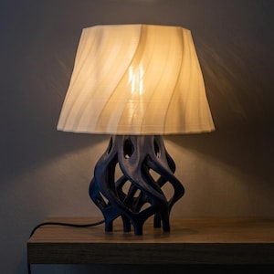 May include: A modern table lamp with a unique design. The lamp features a dark blue, open-work base and a cream-colored, textured lampshade. The shade has a spiral pattern. The lamp is plugged into an outlet.