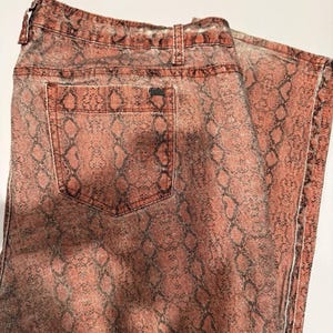 May include: A pair of pants featuring a faded terracotta and black snakeskin print pattern. The garment includes a standard waistband with belt loops, a single visible back pocket, and reinforced stitching along the seams and pocket edges.