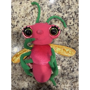 Op de afbeelding: Een pluche insect met een roze lichaam, groene antennes en ledematen en grote, expressieve ogen. Het speelgoed heeft gouden vleugels en is ontworpen om op een vriendelijke kever te lijken. Het speelgoed is zacht en knuffelig, perfect voor kinderen.