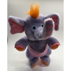 May include: A plush purple elephant toy with orange hair, blue eyes, and pink accents on the ears, trunk, and feet. The elephant wears a pink and purple striped diaper. A cute and cuddly stuffed animal.