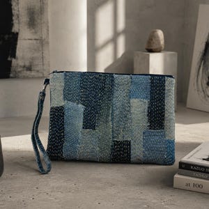 May include: A rectangular zippered clutch bag featuring a patchwork design of various blue denim fabric strips. The exterior is decorated with white running stitch embroidery throughout. A matching denim wrist strap is attached to the side zipper.
