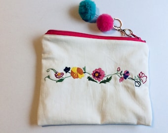 May include: White canvas zippered pouch with a pink zipper and embroidered flowers in shades of pink, yellow, orange, and green. The pouch has a pink and blue pom-pom attached to a gold key ring.