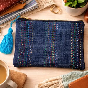 Può includere: Una pochette in denim con cuciture verticali colorate e un nappa blu. La borsa rettangolare ha una cerniera dorata e uno sfondo blu scuro. Le cuciture includono fili rossi, gialli, verdi e blu. La pochette è su una superficie di legno chiaro.