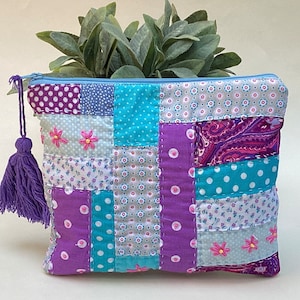 Op de afbeelding: Een patchwork-ritszakje met een paarse kwast. Het zakje is gemaakt van verschillende stoffen in tinten paars, blauw en wit met roze bloemenborduursel.