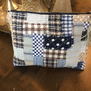 Puede incluir: Una bolsa de cremallera de tela patchwork con un borla marrón. La bolsa está hecha de varias telas azules, blancas y marrones con diferentes patrones, incluyendo cuadros, lunares y a cuadros.
