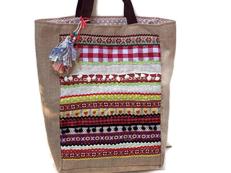 May include: A brown burlap tote bag with a colorful patchwork design. The design features red and white gingham, green and yellow stripes, and white pom-poms.