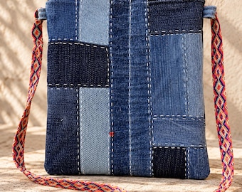 May include: A rectangular crossbody bag constructed from patchwork denim fabric in various shades of blue, featuring visible white running stitch detailing and a single red stitch accent. The bag has a black zipper and a colorful woven strap.