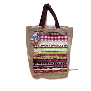 May include: A brown jute tote bag with a colorful patchwork design. The bag features a variety of ribbons, trims, and fabrics in red, green, yellow, and white. The bag has brown handles and a small tassel.