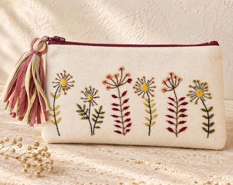 May include: A rectangular cream-colored canvas pouch featuring an embroidered design of six wildflowers in shades of yellow, green, and maroon. The pouch has a dark maroon zipper closure and a multi-colored fabric tassel attached to the side.