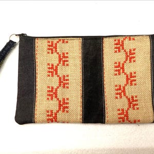 Könnte beinhalten: Eine rechteckige Reißverschlusstasche mit einem beigen Jute-Mittelteil, das ein rotes Kreuzstichmuster aufweist, flankiert von dunklen Jeansstoff-Abschnitten und einem zentralen schwarzen Samtstreifen. Eine passende Handschlaufe aus Jeansstoff ist befestigt.