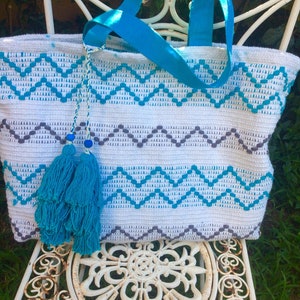 Pode incluir: Sacola tote de croch&ecirc; branca com padr&atilde;o chevron azul e cinza. A sacola tem borlas azuis e uma al&ccedil;a de fita azul.