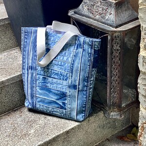 May include: A blue denim tote bag with white straps. The bag is made from recycled denim and has a striped pattern. The bag is sitting on a set of concrete steps.
