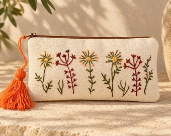 May include: A rectangular off-white canvas pouch featuring colorful floral embroidery in shades of yellow, orange, and dark red. The pouch has a brown zipper closure and a bright orange tassel attached to the zipper pull, adding a vibrant accent.