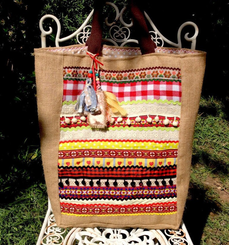 May include: A large, brown burlap tote bag with a colorful patchwork design. The bag is decorated with red and white gingham, yellow, red, and green ribbons, and white lace. The bag has a brown handle and a tassel.