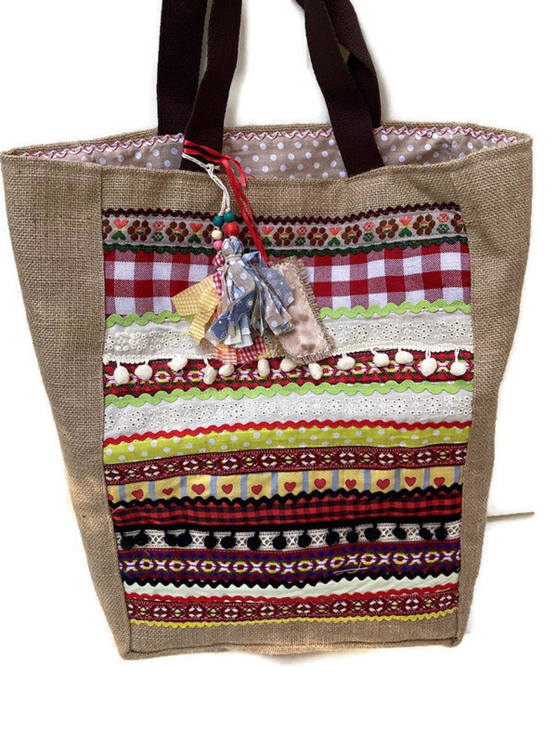 May include: A large, brown burlap tote bag with a patchwork design. The bag features a variety of colorful fabrics, including red and white gingham, yellow and green stripes, and floral patterns. The bag has brown fabric handles and a tassel charm.