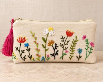 May include: A rectangular beige canvas pouch featuring colorful floral embroidery across the front. The design includes various stems, leaves, and blossoms in red, blue, yellow, and pink. A bright pink tassel is attached to the gold zipper pull.