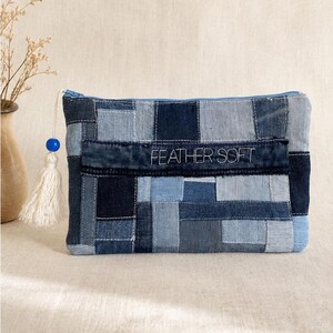 May include: A rectangular zippered pouch crafted from a patchwork of various blue denim fabric squares. The front features a horizontal strip with the embroidered text FEATHER SOFT. A white tassel with a blue bead is attached to the zipper pull.