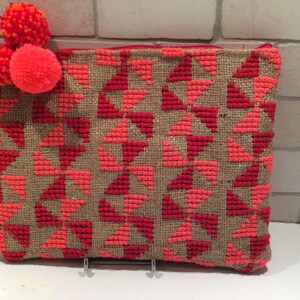 May include: A brown and red woven clutch with a geometric pattern. The clutch has a zipper closure and is decorated with three red pom-poms.