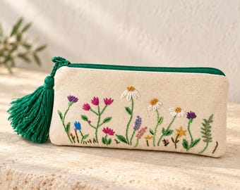 May include: A rectangular beige canvas pencil case featuring colorful floral embroidery of daisies, cornflowers, and wildflowers. The case has a dark green zipper closure and a matching dark green decorative tassel attached to the side zipper pull.