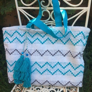 Pode incluir: Sacola tote de croch&ecirc; branca com um padr&atilde;o chevron turquesa e cinza. A bolsa tem uma al&ccedil;a de tecido turquesa e um tassel turquesa.