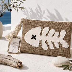 May include: A rectangular brown burlap zipper pouch featuring a large white fabric fish skeleton appliqué with a black stitched X for an eye. A beige tassel is attached to the zipper pull, along with a small rectangular tag that reads thank you for shopping handmade by APOPSIS.