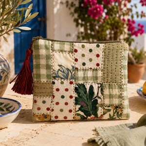 Puede incluir: Un neceser con cremallera de patchwork con una borla burdeos. El neceser presenta una mezcla de cuadrados de tela de cuadros verdes, lunares y estampados florales. El fondo incluye un jarrón con ramas de olivo, un cuenco pequeño y limones.