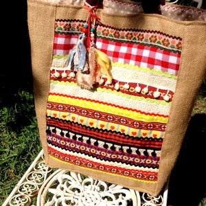 May include: A large, brown burlap tote bag with a colorful patchwork design. The bag features red, yellow, white, and black stripes, along with a red and white checkered pattern. The bag has a tassel with beads and pom-poms.