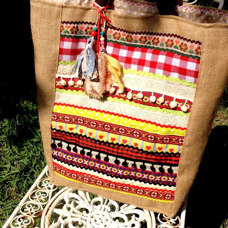 May include: A large, brown burlap tote bag with a colorful patchwork design. The bag features red, yellow, white, and black stripes, along with a red and white checkered pattern. The bag has a tassel with beads and pom-poms.