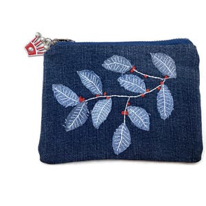 May include: A small blue denim zippered pouch with a white embroidered design of leaves and red berries. The zipper pull has a small crown charm.
