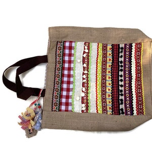 May include: A brown burlap tote bag with a colorful striped design. The bag has a brown fabric handle and a tassel made of colorful ribbons.