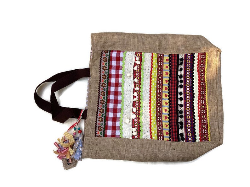 May include: A brown burlap tote bag with a colorful striped design. The bag has a brown fabric handle and a tassel made of colorful ribbons.