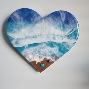 May include: Heart-shaped resin art piece depicting an ocean scene. The artwork features shades of blue and white, mimicking waves. Small seashells and a starfish are embedded on a wooden base, creating a beach effect.