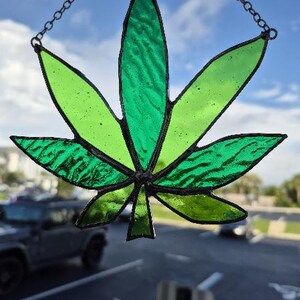 Könnte beinhalten: Ein Cannabisblatt aus Glas in Grüntönen mit schwarzen Konturen und einer Kette zum Aufhängen. Das Blatt hängt vor einem blauen Himmel und einem Parkplatz.