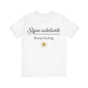 Puede incluir: Camiseta blanca con las palabras "Sigue adelante" sobre "Keep Going" en escritura negra. Un sol dorado está debajo del texto. Es una prenda de vestir informal.