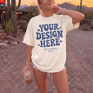 May include: An off-white short-sleeve t-shirt featuring the dark blue text YOUR DESIGN HERE in a retro-style font, accented with small heart icons. The shirt includes the small text THELOVEMOCKS and a row of three tiny hearts below the main graphic.
