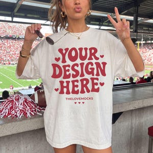 Może przedstawiać: Biały t-shirt z krótkim rękawem z czerwonym napisem YOUR DESIGN HERE, ozdobiony małymi czerwonymi sercami. Koszulka zawiera również napis THELOVEMOCKS z trzema małymi czerwonymi sercami pod główną grafiką.