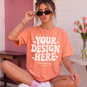 May include: Peach-colored t-shirt with the text "YOUR DESIGN HERE" in white, with heart accents. The text "THELOVEMOCKS" is below the main text. The shirt is short-sleeved and appears to be made of a soft material.