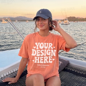 May include: An orange t-shirt with the text "YOUR DESIGN HERE" in white, with heart accents. The model is wearing a navy blue baseball cap and gold hoop earrings. The background shows a body of water and boats.