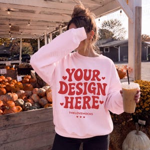 May include: A light pink crewneck sweatshirt featuring the red text YOUR DESIGN HERE with small heart accents and the text THELOVEMOCKS printed below. The sweatshirt has long sleeves and a ribbed collar, cuffs, and hem, worn over dark pants.