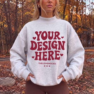 May include: A light gray heathered sweatshirt with a mock neck and long sleeves. The front features bold, dark red text that reads "YOUR DESIGN HERE" surrounded by small heart icons. Below the main text, the word "THELOVEMOCKS" is printed.