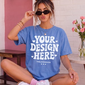 May include: A blue t-shirt with the text "YOUR DESIGN HERE" in white, with the words "THELOVEMOCKS" below. The model is wearing sunglasses and jewellery.