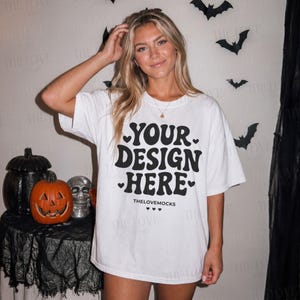 Puede incluir: Una camiseta blanca de manga corta con texto negro que dice YOUR DESIGN HERE y THELOVEMOCKS junto a pequeños iconos de corazones. La prenda cuenta con cuello redondo, un corte holgado y una tipografía ondulada en la parte frontal.