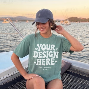 Könnte beinhalten: Ein salbeigrünes T-Shirt mit dem Text "YOUR DESIGN HERE" in Weiß, mit Herzakzenten. Das Model trägt eine marineblaue Baseballkappe. Der Hintergrund zeigt ein Gewässer und Boote.