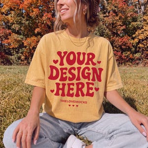 May include: A mustard yellow short-sleeve t-shirt featuring the red text YOUR DESIGN HERE in a bold, wavy font. The design includes small red heart icons surrounding the text and the smaller red text THELOVEMOCKS with three hearts below it.