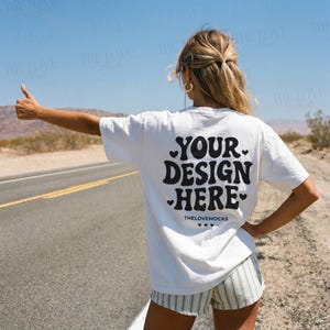 Può includere: T-shirt bianca con la scritta nera "YOUR DESIGN HERE", con un font retrò e cuori. La maglietta viene indossata all'aperto in una giornata di sole. Il testo "THELOVEMOCKS" è sotto.