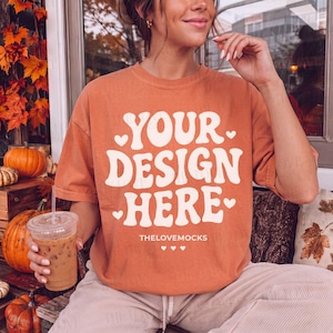 May include: A burnt orange short-sleeve t-shirt featuring the white text "YOUR DESIGN HERE" in a bold, wavy font, accented with small white hearts. Below the text, the words "THELOVEMOCKS" appear in a smaller font with three tiny hearts.
