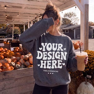 May include: A dark gray long-sleeve sweatshirt featuring white text that reads YOUR DESIGN HERE and THELOVEMOCKS with small heart icons. The sweatshirt has a crew neckline and ribbed cuffs, displayed in a setting with pumpkins and a drink.